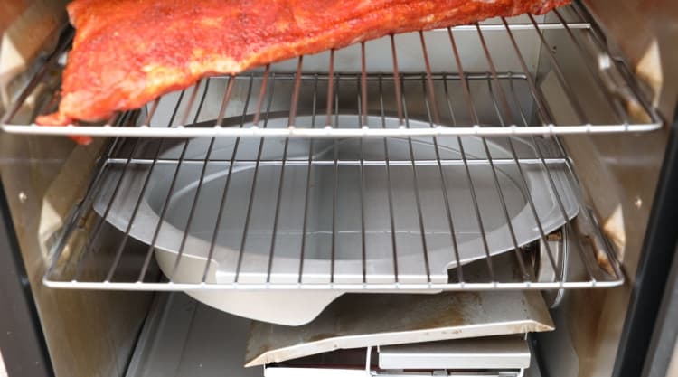 A rack of ribs on top shelf of an electric smoker with a water pan below