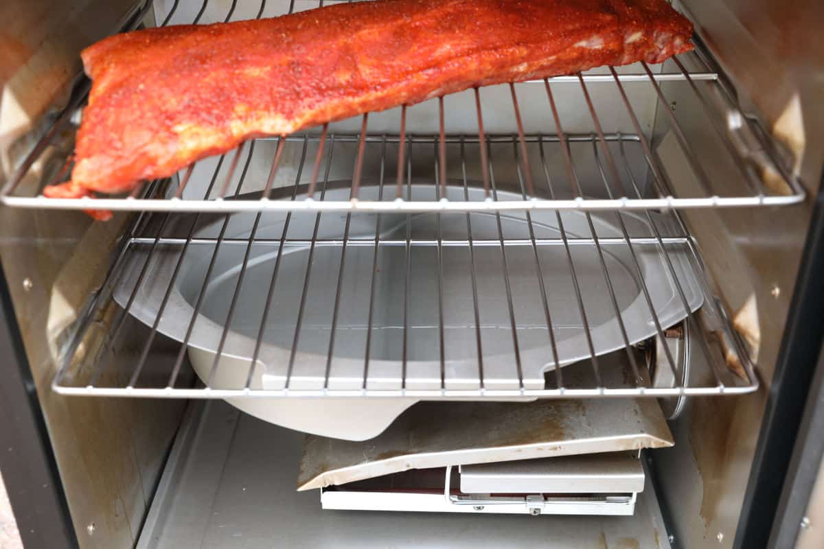 A rack of ribs on top shelf of an electric smoker with a water pan below