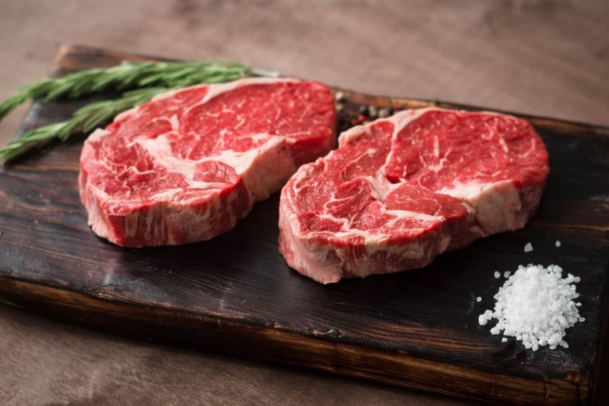 Two 1 inch thick ribeye steaks on a cutting board with rosemary and a pile of sea s.