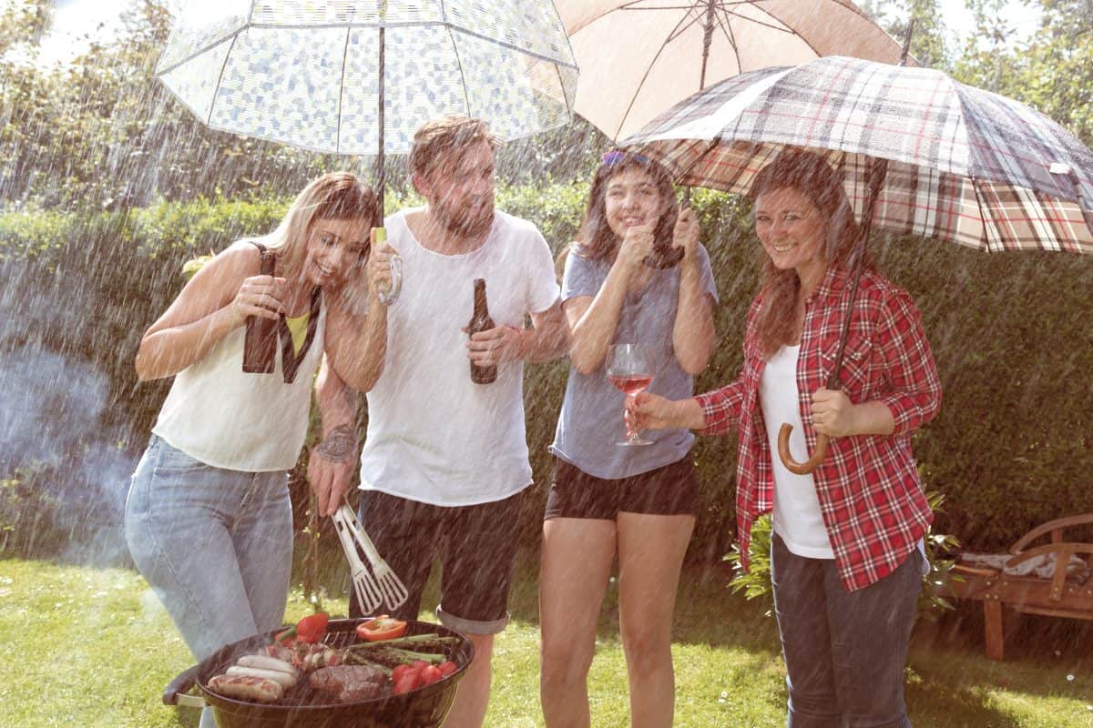 A young crowd of 4 under umbrellas having a BBQ in the r.
