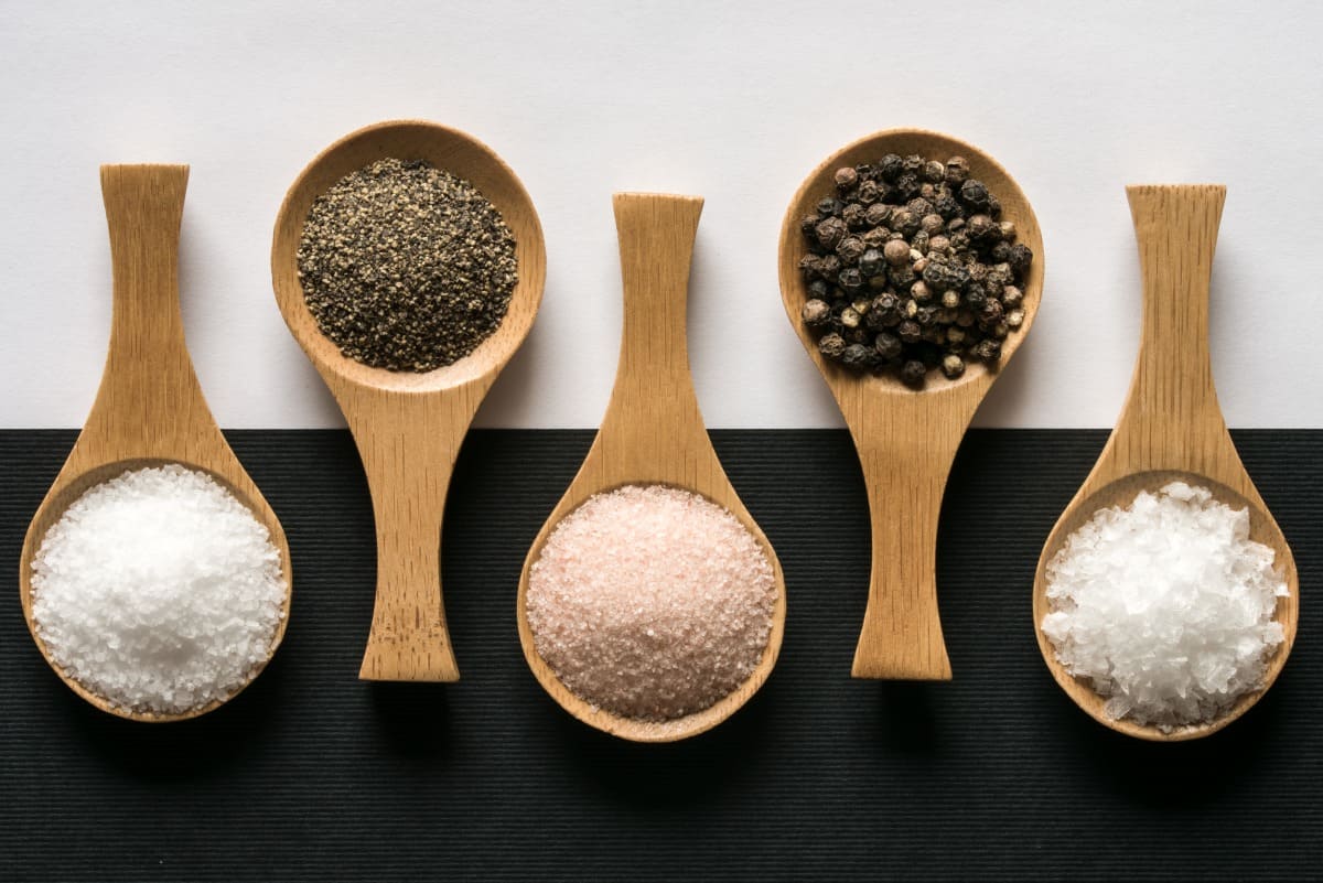 Kosher salt, ground pepper, himalayan salt, peppercorns, and sea salt flakes on wooden spoons on a black and white backgro.