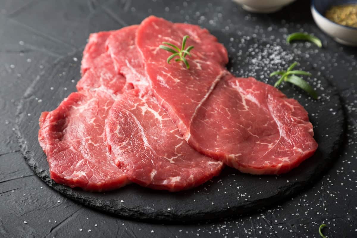 Three steaks under 1 inch thick on a dark cutting board with some rosemary and spi.