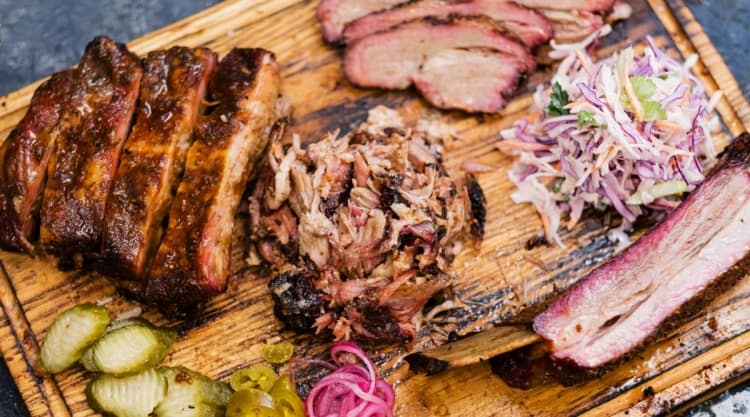 Different smoked meats, slaw and pickles as a BBQ platter on a cutting board.
