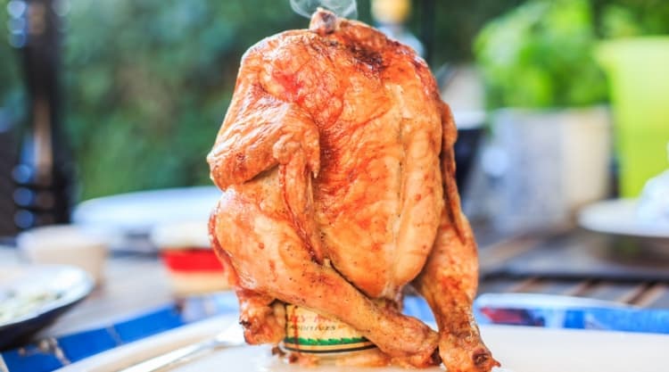 beer can chicken, grilled, sitting upright on a white cutting board with a blurred garden background.