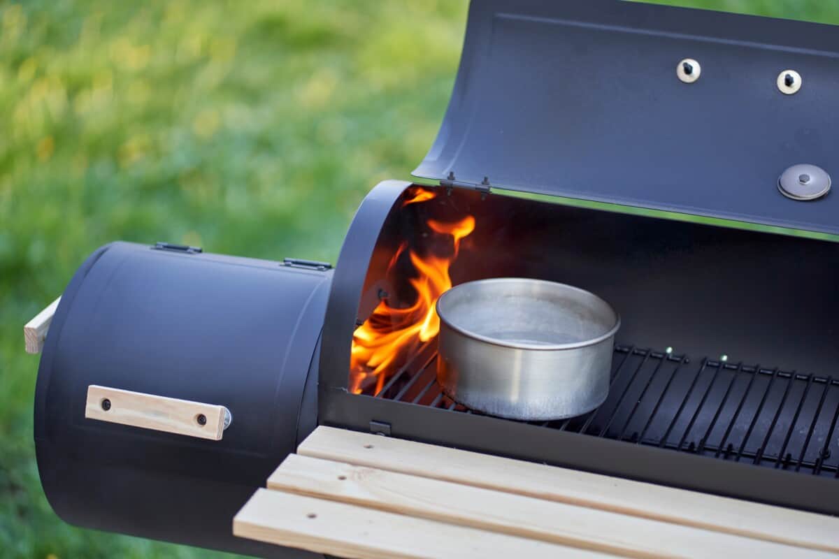 An offset smoker with lid open, showing flames licking out of the firebox, and a pan of water on the grates.