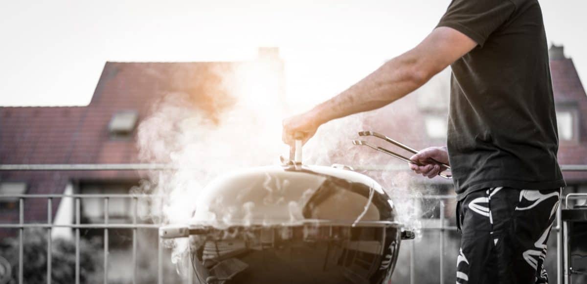 A man holding the lid on a smoking charcoal gr.