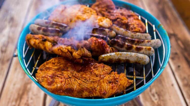 A blue charcoal grill being used on a wooden deck loaded with meats grilling.