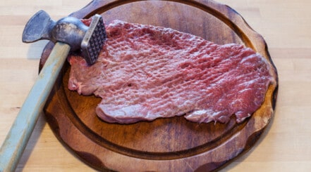 A piece of meat being tenderized by a mallet while on a round chopping board.