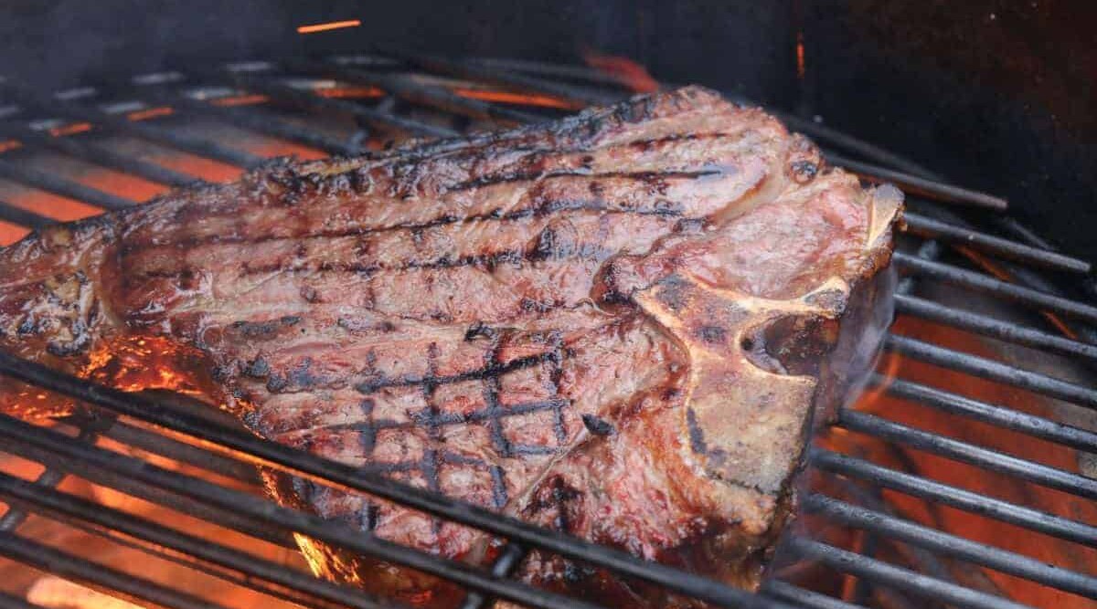 Thick porterhouse steak being seared on a charcoal grill.
