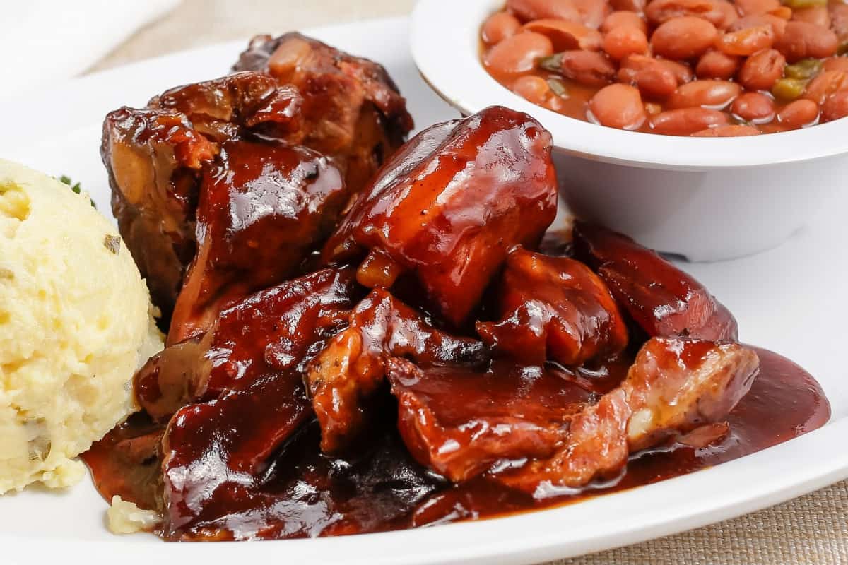 rib tips glazed with BBQ sauce, on a white plate with mashed potato and some beans.