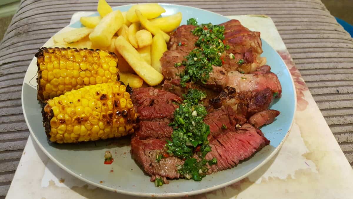 Mixed grilled meats, sweetcorn, and chips on a dinner plate.