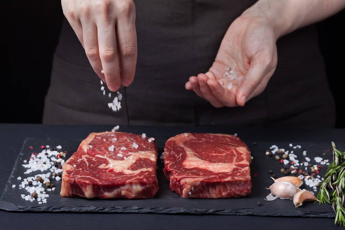 A woman's hands sprinkling salt onto two steaks on a slate surf.