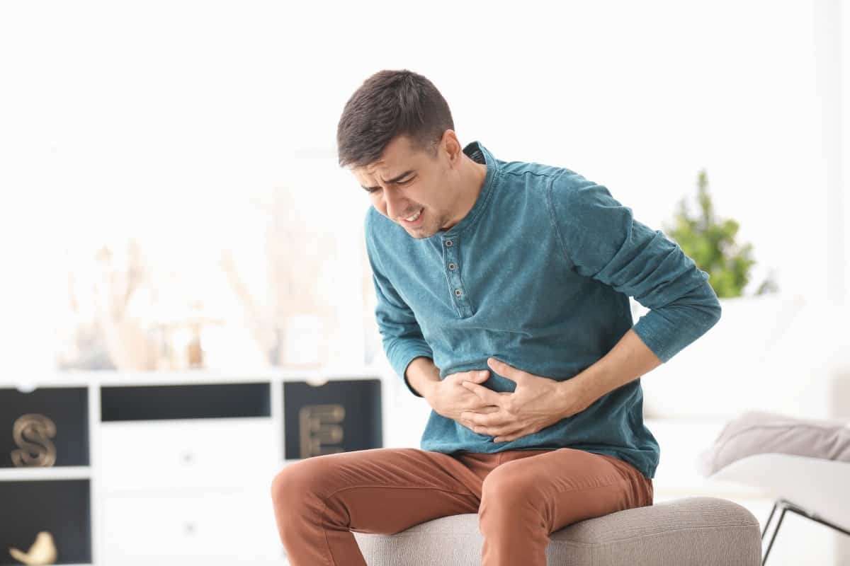 A man in brown jeans and blue shirt, sitting down holding his stomach looking s.