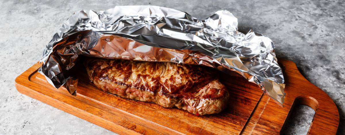 A grilled steak resting on cutting board under loosely tented f.