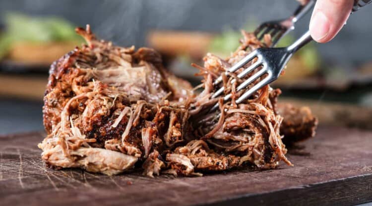 Chunk of BBQ smoked pork being pulled apart by 2 forks on a wooden chopping board