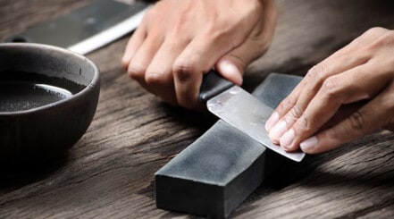 A knife being sharpened on a whetstone next to bowl of water on wooden table.