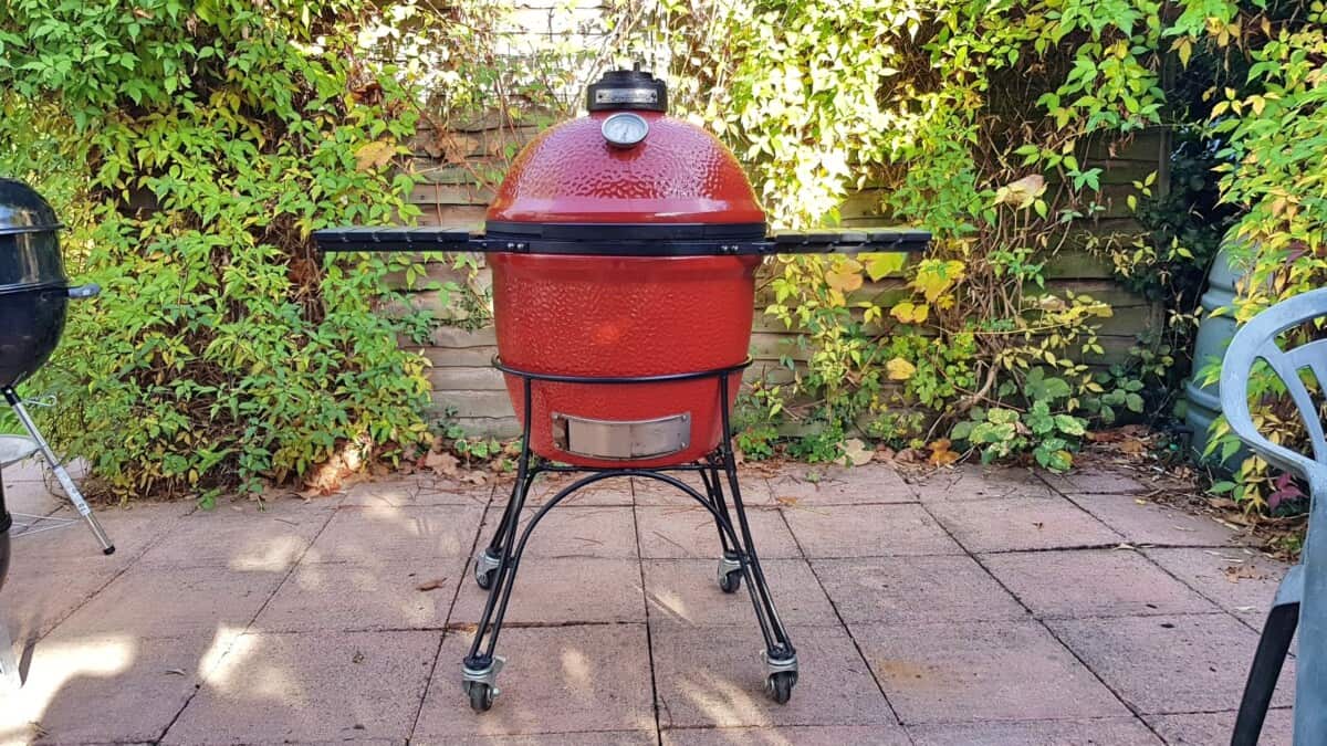 A red Kamado Joe Classic 1 on a paved patio, with a fence and bushes behind it.