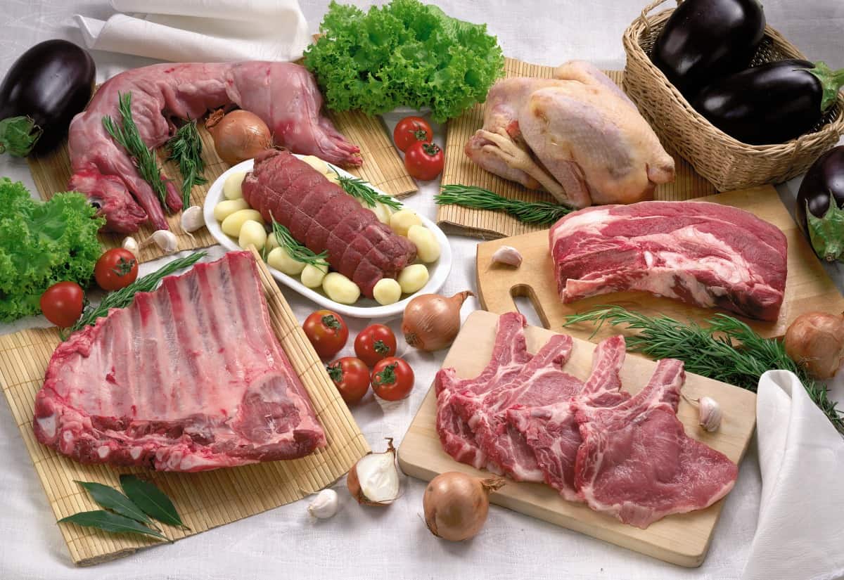 A selecton of raw meat and poultry on cutting boards, with veg on a white backgro.