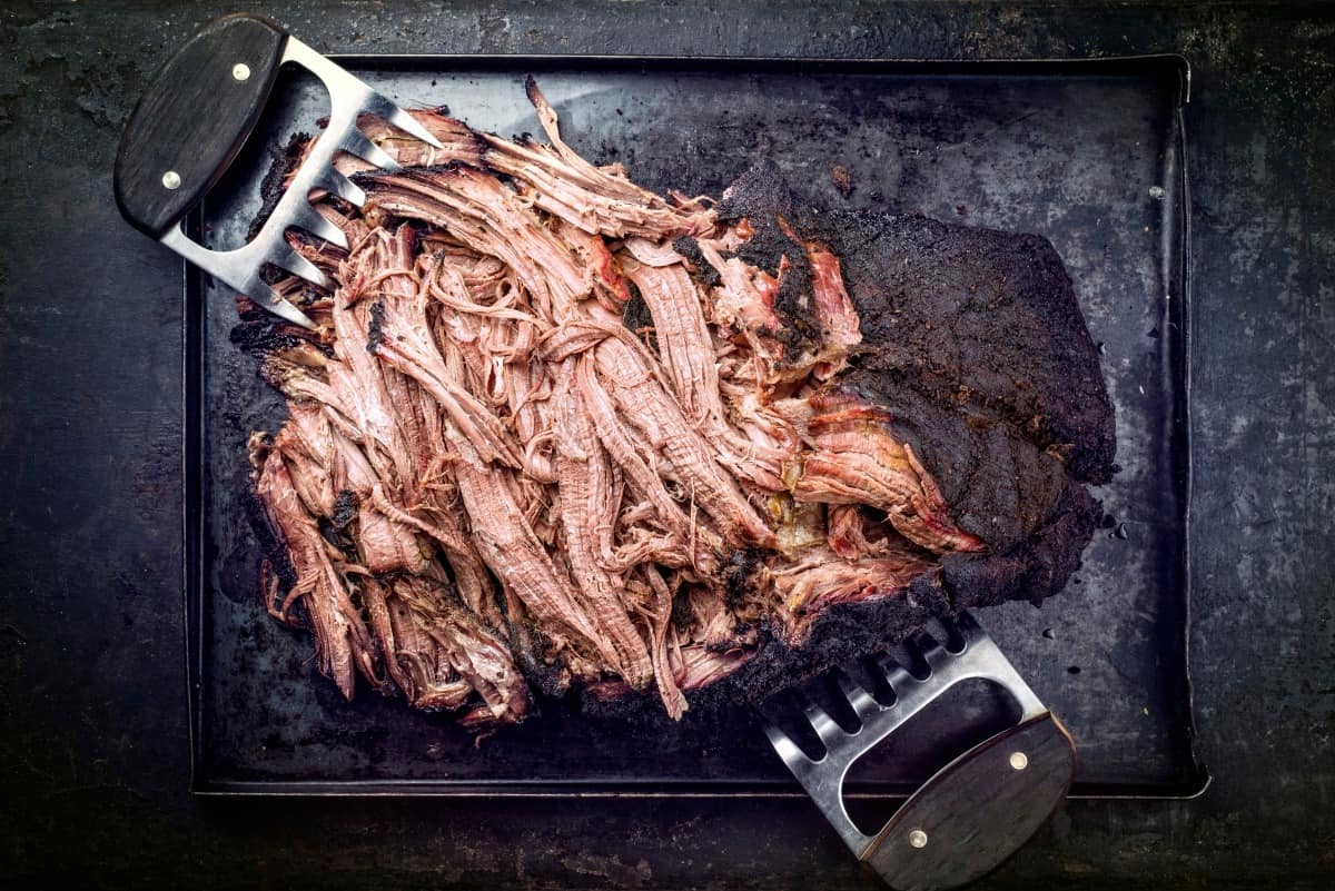 Overhead shot of some BBQ pulled pork in a serving tray, on rustic board, with meat cl.