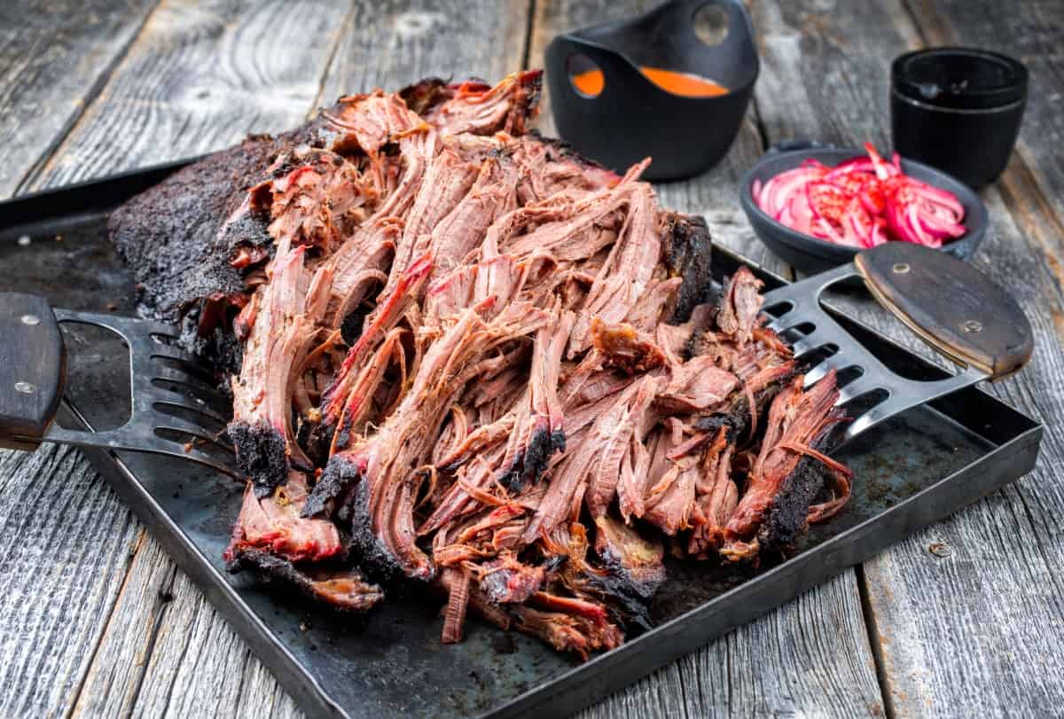 pulling pork with claws in a large metal tray, with red slaw and sauce out of focus in th ebackgro.