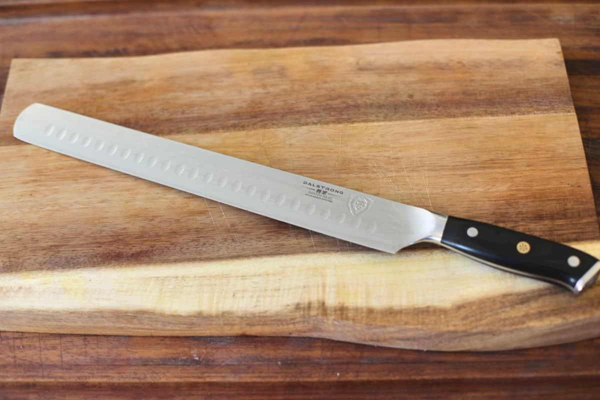 a large, Dalstrong scalloped slicing knife on a wooden chopping bo.