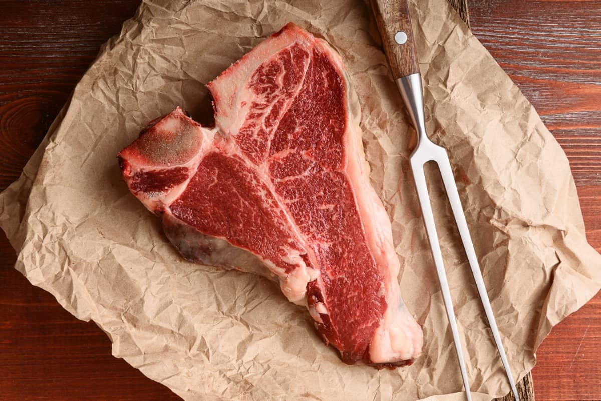 A raw t-bone steak resting on steak paper with a 2-pronged f.