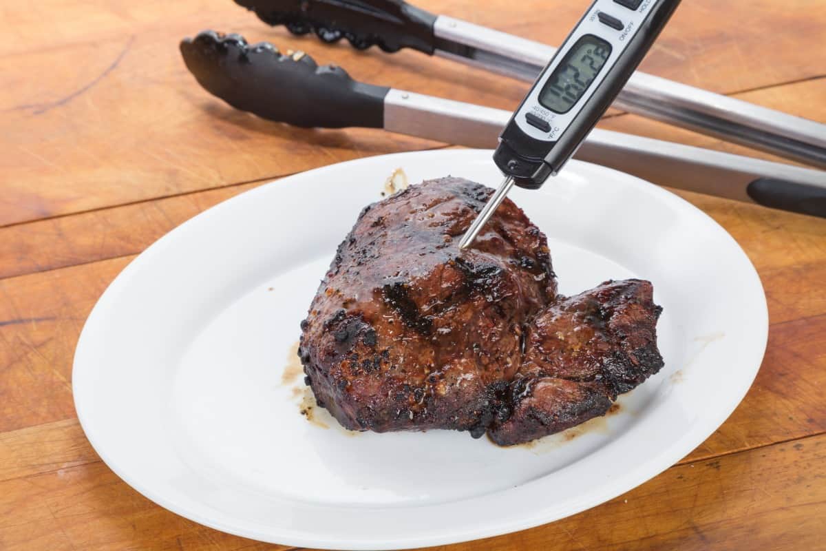 An instant read thermometer measuring temp of some steak on a pl.