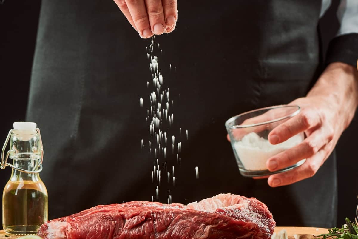 A man sprinkling salt onto a steak, on a cutting board, next to an oil bottle and a pepper grin.