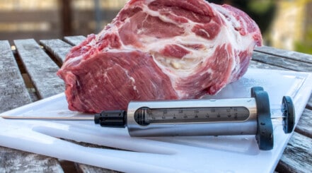 A meat injector sitting on a white chopping board beside a large joint of pork.