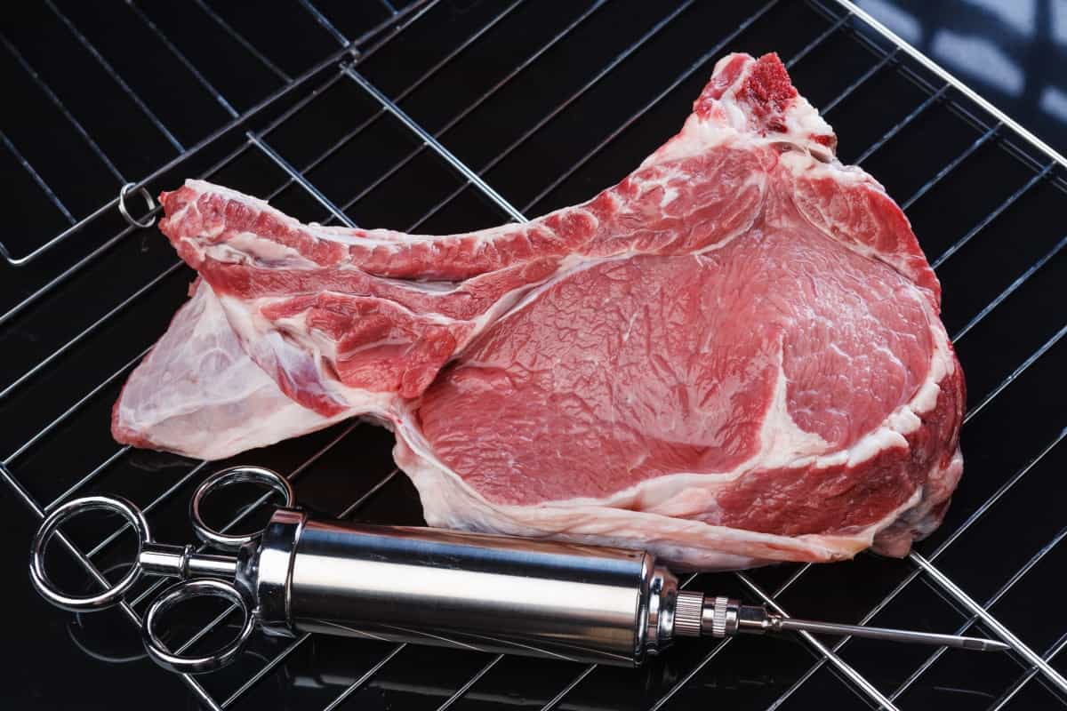 A metal marinade syringe sitting next to some beef on a grill r.
