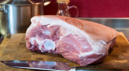 Close up of a pork butt, fat side up, on a wooden chopping board with a kitchen knife in.