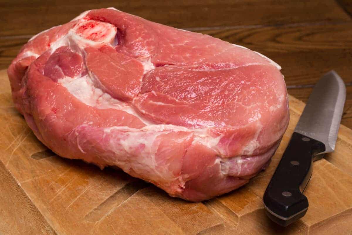 a cut of pork shoulder on a wooden chopping board, with a kitchen knife next to.