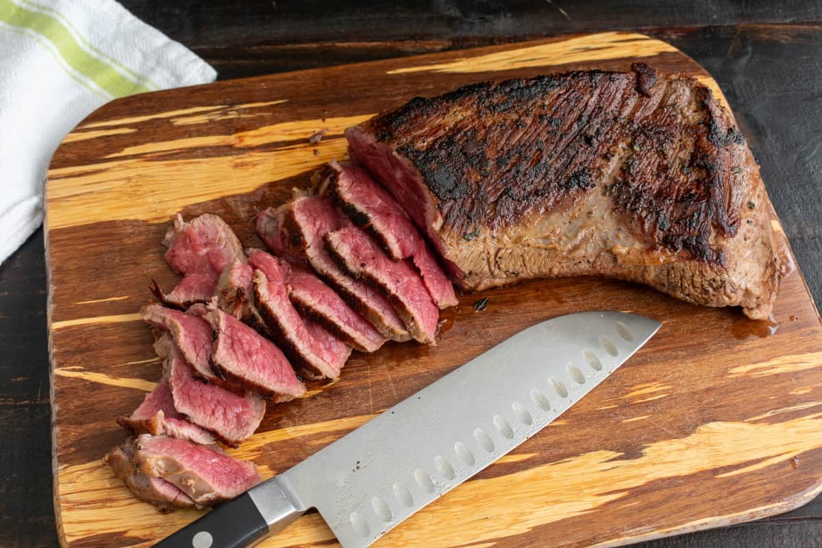 A rare grilled tri-tip, sliced to halfway through the roast, sitting on a bamboo cutting bo.