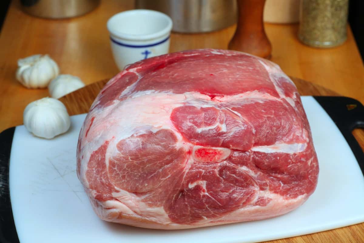 A raw bone in pork shoulder on a white cutting board with garlic bu.