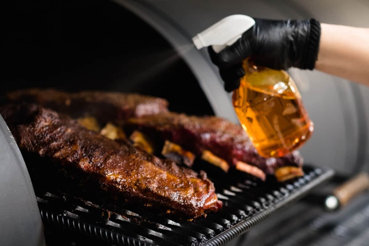 Black gloved hands spritzing ribs in a large smoker