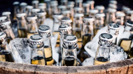 Close up of the top of lots of bottles of beer in an outdoor cooler in ice.