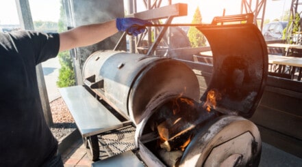 Large BBQ smoker with fire box held open by a man with a gloved hand.