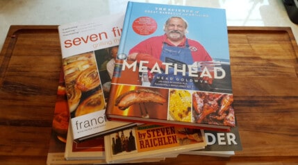 Some of the best BBQ and grilling cookbooks piled on each other on a table.