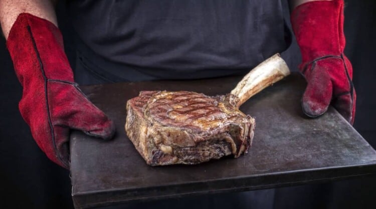 Man with red leather grilling gloves carrying a tomahawk steak on a slate board.