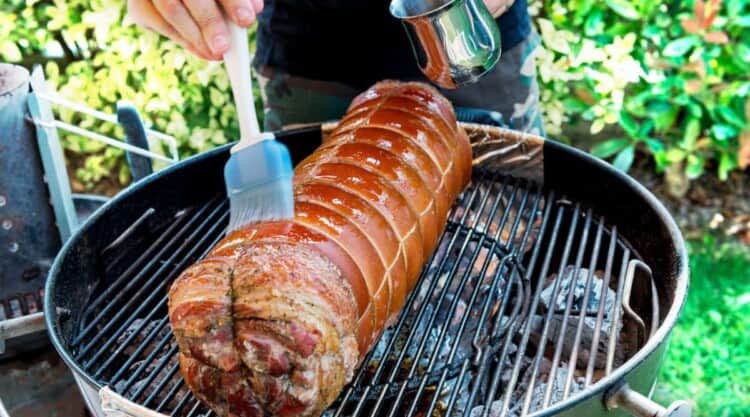 BBQ mop sauce being smeared all over a porchetta on a grill.