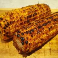 2 grilled corn on the cob without husks, sitting on a wooden board.