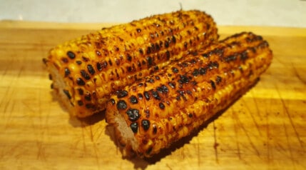 2 grilled corn on the cob without husks, sitting on a wooden board.
