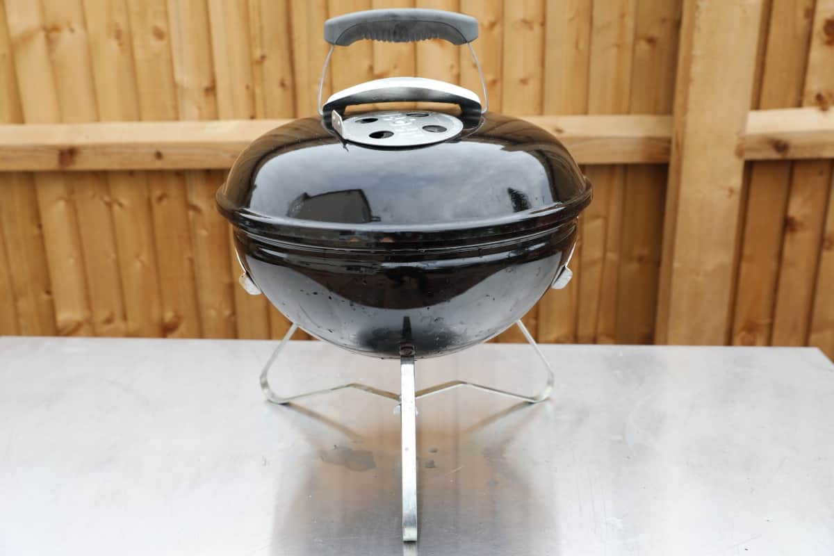  Weber smokey joe on a stainless steel table in front of a wooden fe.