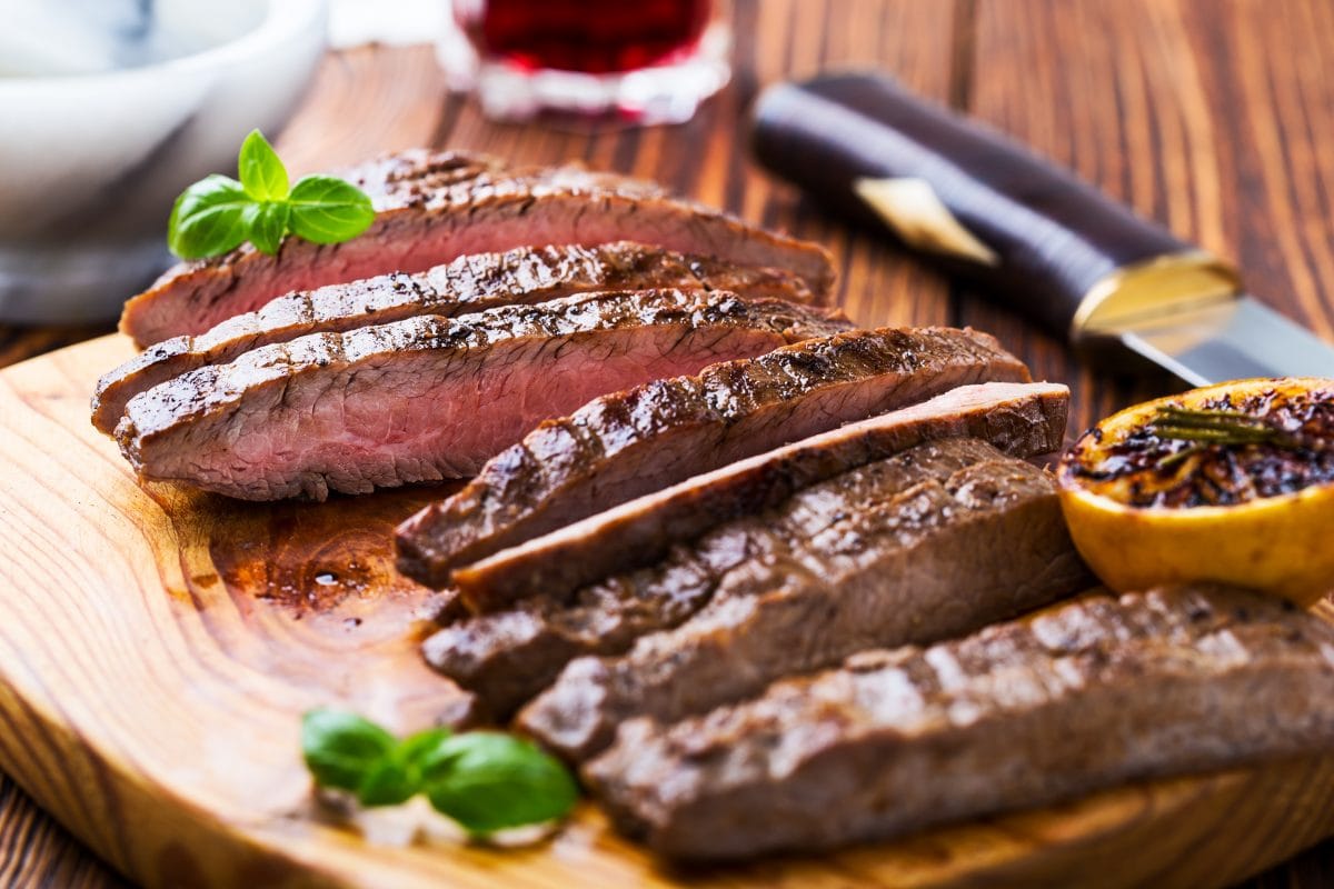 Sliced flank steak with a grilled lemon, and some herbs on a cutting bo.