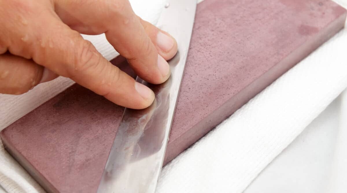 Mans hands sharpening a kitchen knife on a purple whetstone resting on a white towel.