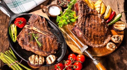 A porterhouse and ribeye steak on a cutting board, with vine tomatoes, greens, and salt.