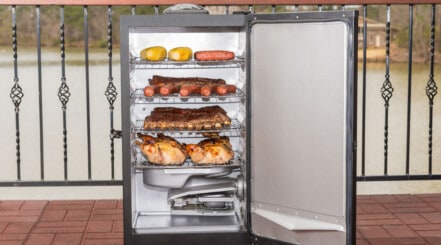 An electric smoker on a wooden balcony, door open and stuffed with food to smoke.