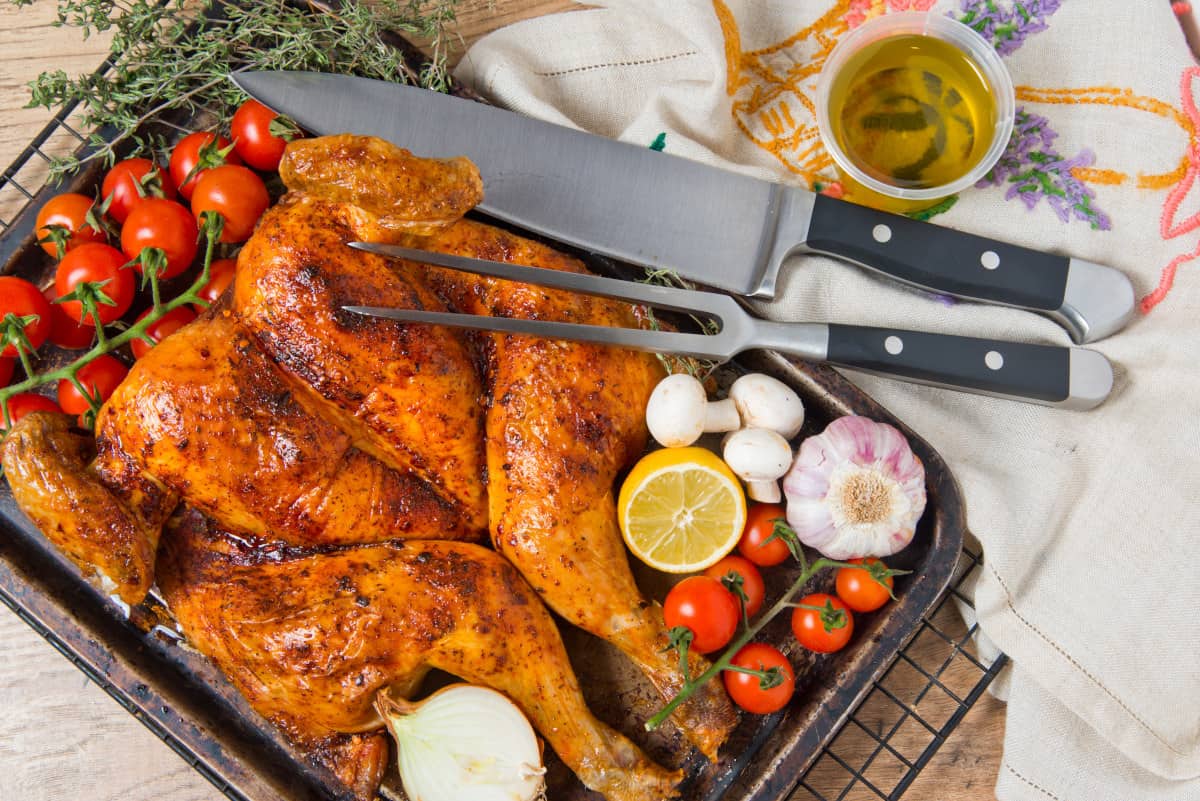 Spatchcock chicken on a tray with onions, garlic, tomatoes, and a carving knife and fork .