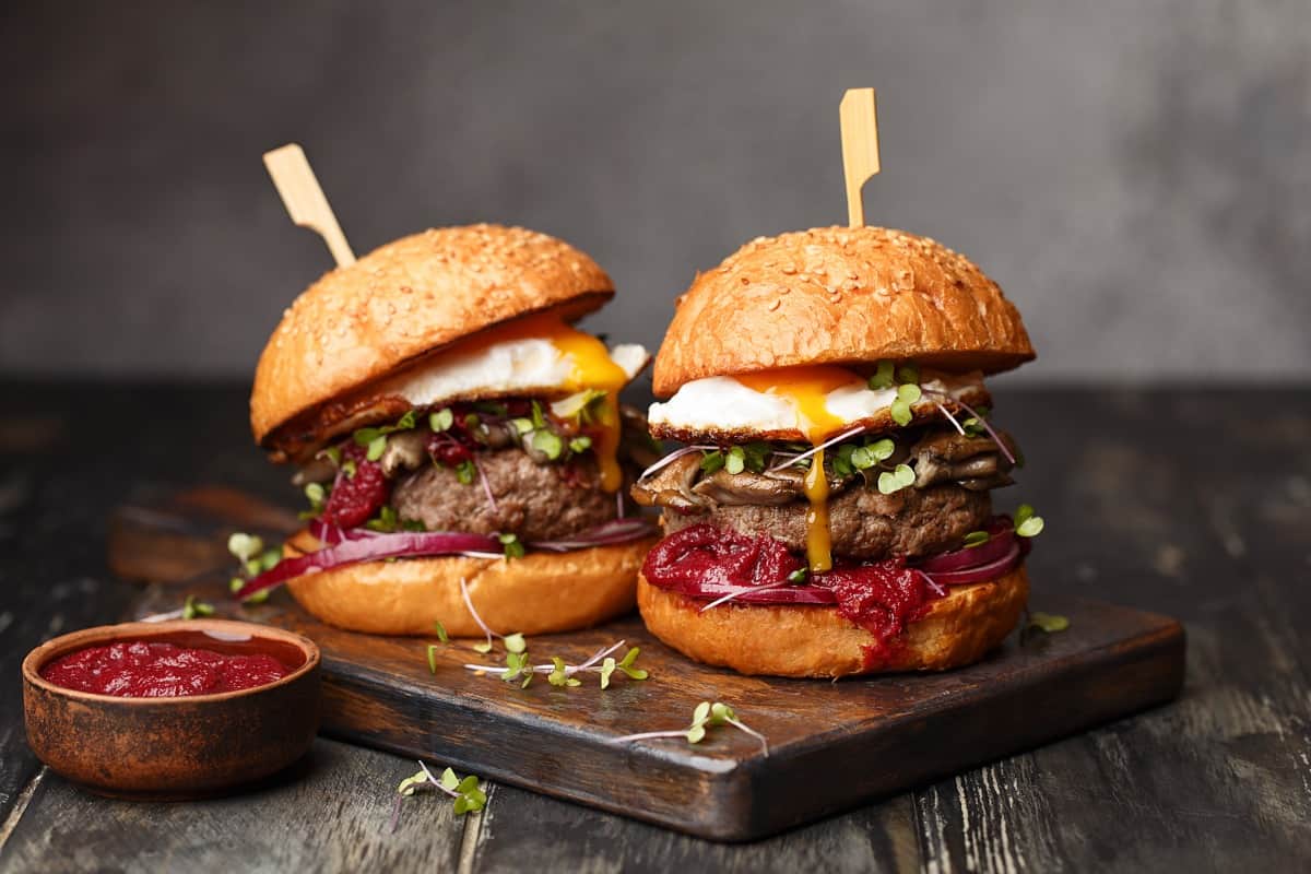 two packed burgers with sticks through them top to bottom, sitting on a wooden cutting bo.