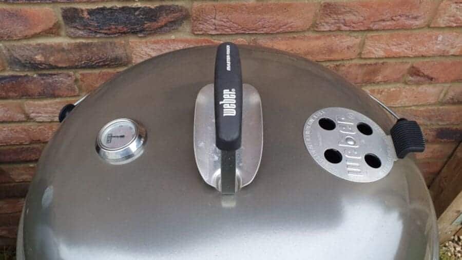 Top view photo of a weber kettle lid, showing thermometer, vent and han.